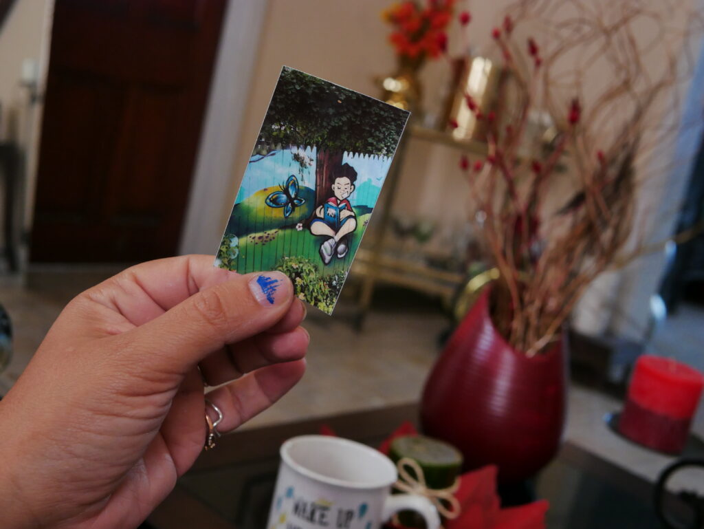 a hand holds a small photograph of an art mural depicting a child sitting under a tree with a book